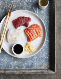 Overhead food photography of assorted sashimi slices with soy sauce and chopsticks on a ceramic plate by Helen Cathcart
