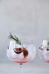 Two glass goblets filled with a pale pink cocktail, ice cubes, sliced figs, dark berries, and a sprig of fresh rosemary against a neutral grey background