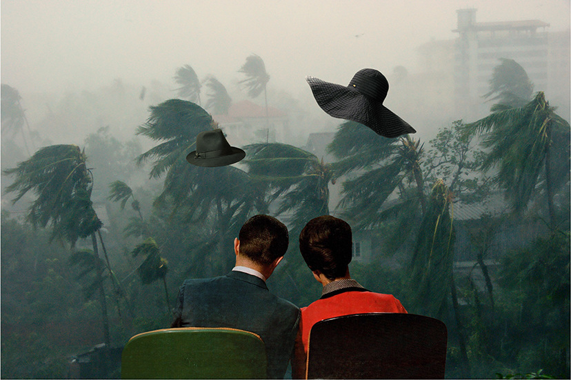 Two people watch from inside as a fierce storm with blowing palm trees rages; two black hats float in the air outside.