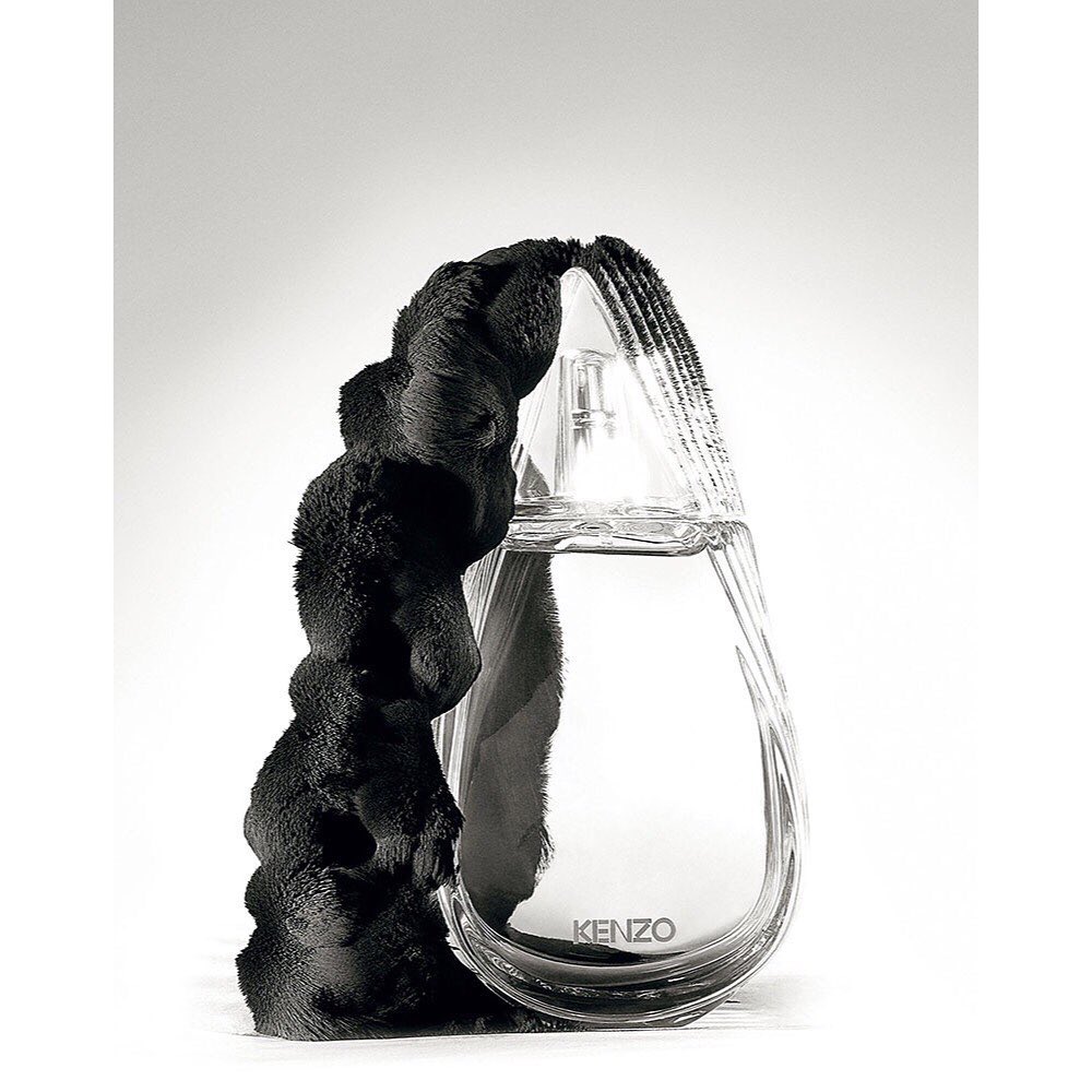Monochrome studio shot of a clear glass KENZO perfume bottle with a black, textured, abstract design element alongside it.