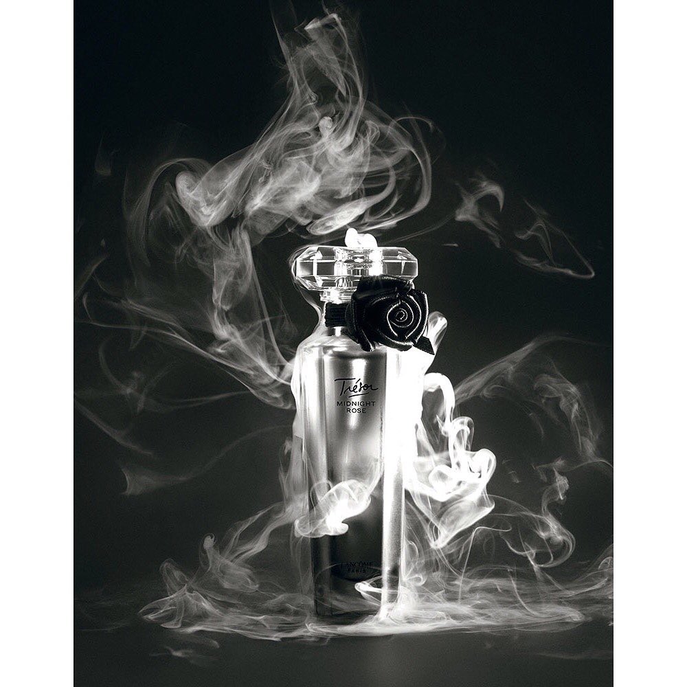 Lancôme Trésor Midnight Rose perfume bottle with black rose detail, surrounded by swirling white smoke against a dark background.