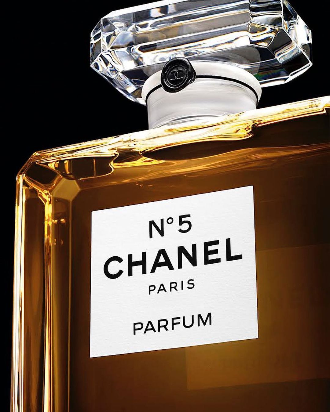 Close-up of a gold-colored Chanel No. 5 Parfum bottle against a black background, featuring a clear geometric cap and white label.