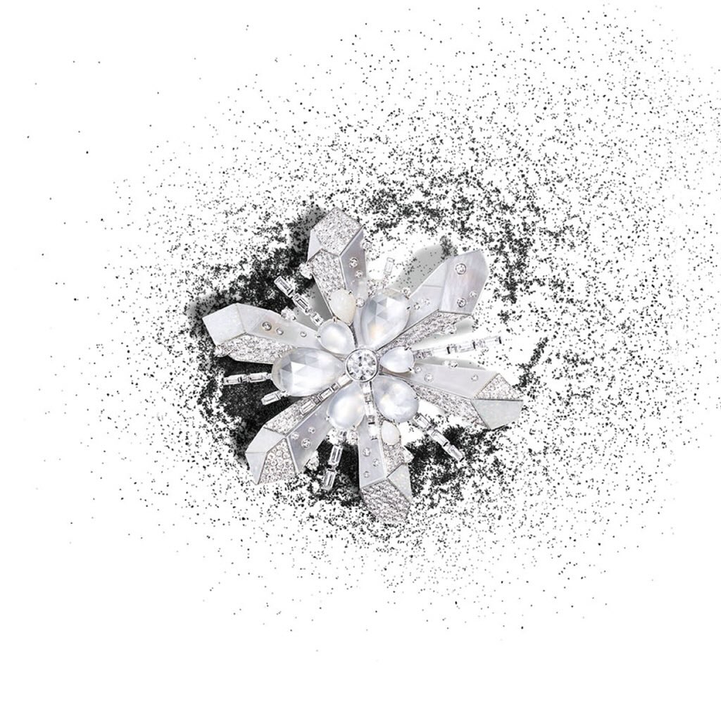 Close-up of a snowflake-shaped brooch made of silver and gems surrounded by black glitter on a white surface, creating a luxurious effect.