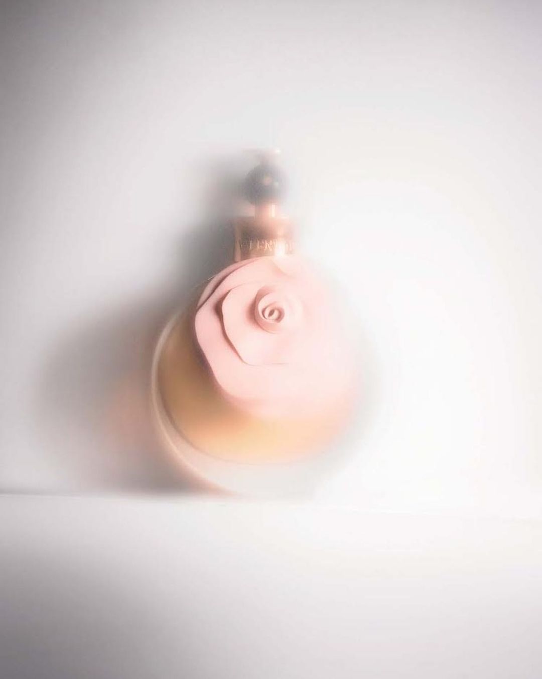 Soft focus shot of a round perfume bottle with a pink flower detail on a white background, creating a delicate and elegant aesthetic.