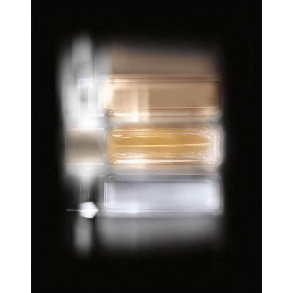 Abstract blurred photograph of three stacked clear cosmetic containers, with silver, gold, and bronze tones, against a dark background.