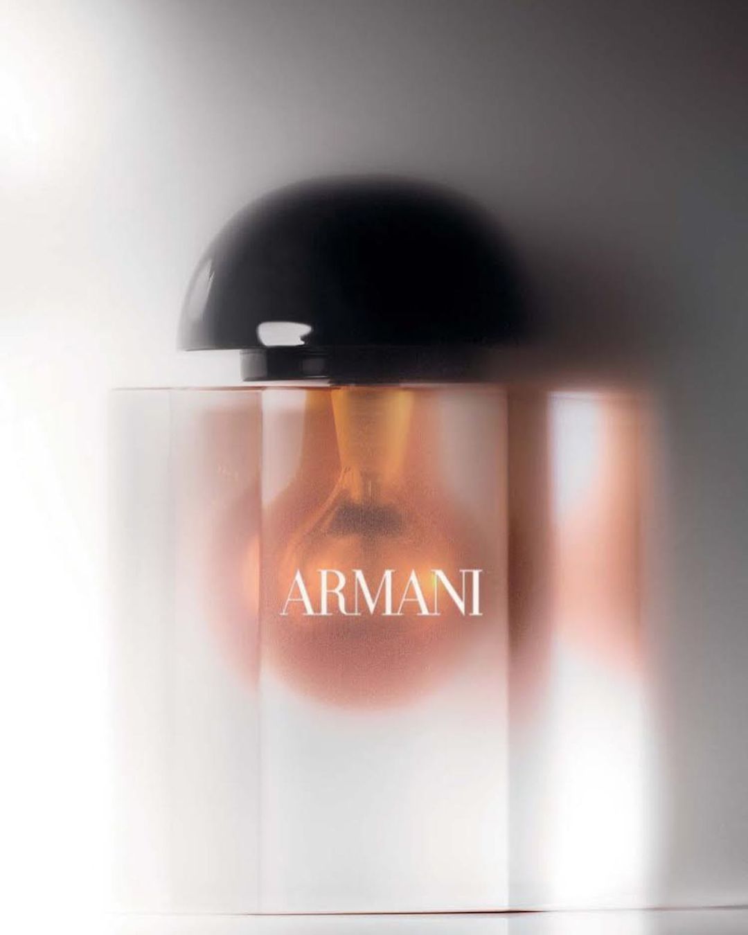 Product shot of a clear glass Armani perfume bottle with a black cap, filled with a light-brown liquid, against a soft gradient background.