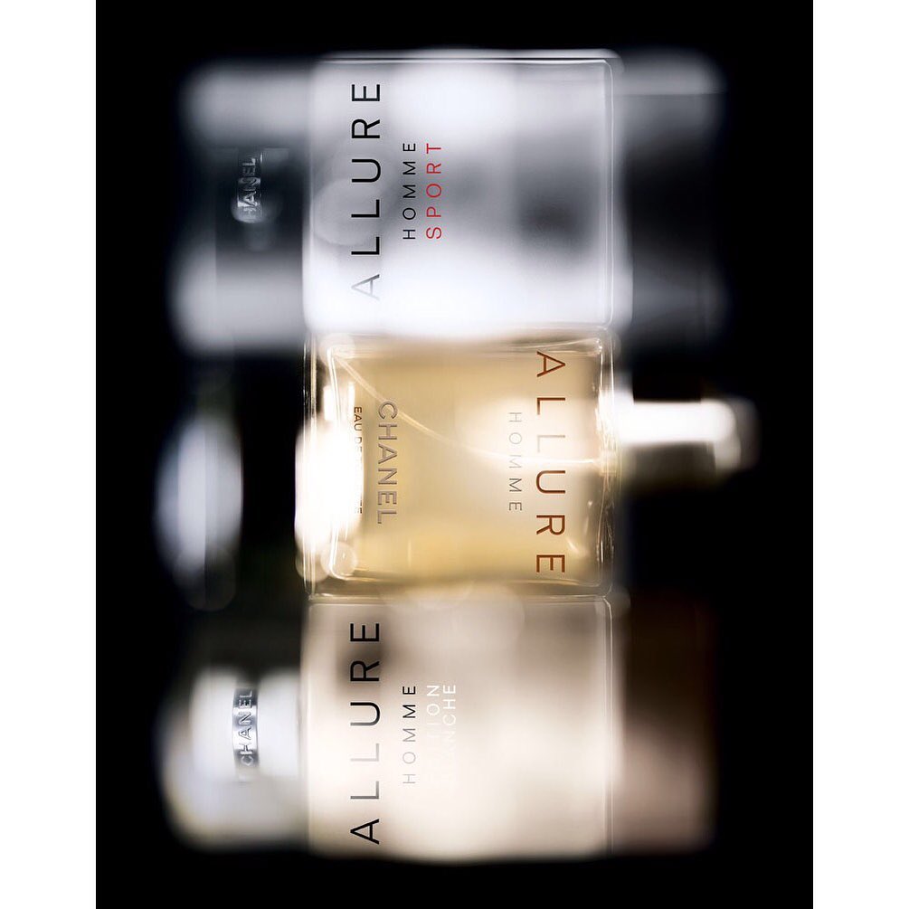 Three Chanel Allure Homme cologne bottles are stacked vertically, showcasing white and gold liquid against a dark, blurred background.