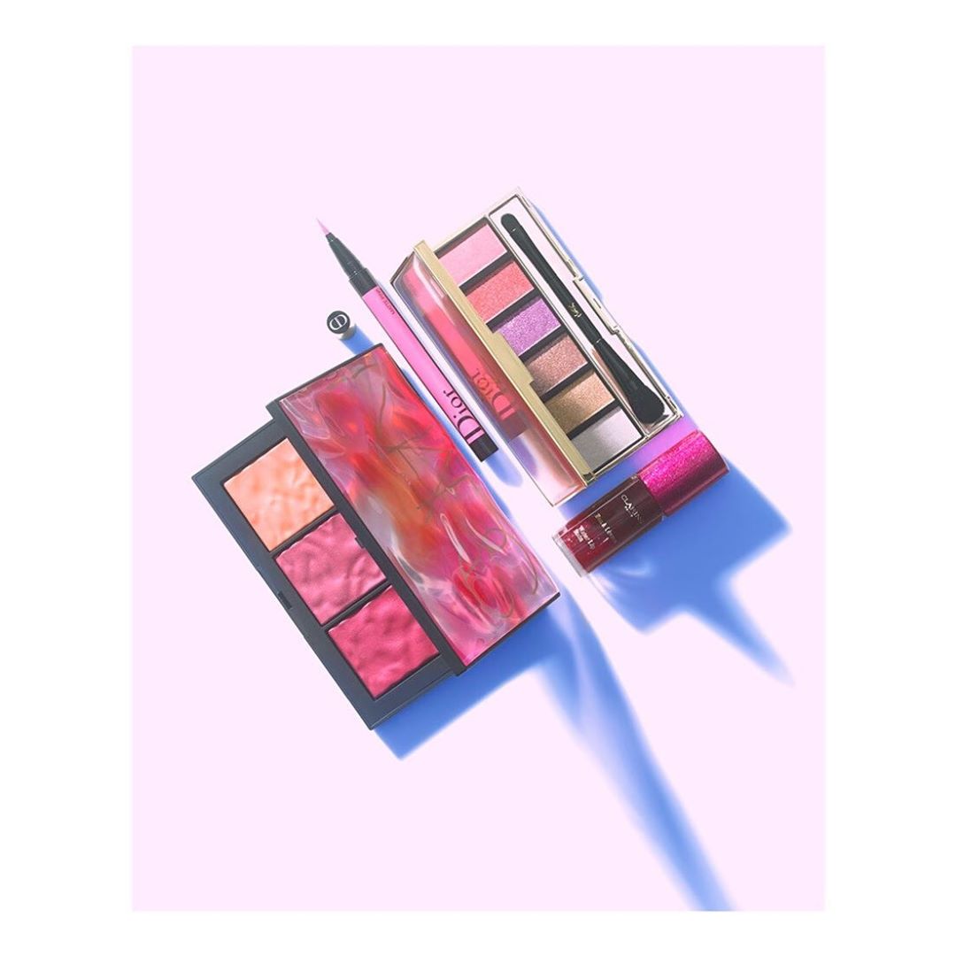 Flat lay of makeup products including a Dior eyeshadow palette, blush palette, eyeliner, and lip gloss on a light purple background.