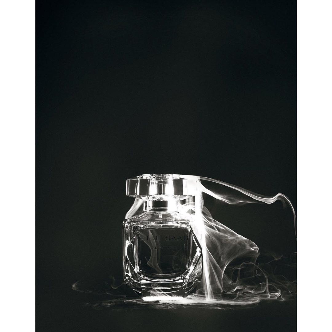 Clear glass perfume bottle with silver accents, surrounded by white smoke against a black background. Minimalist beauty still life.