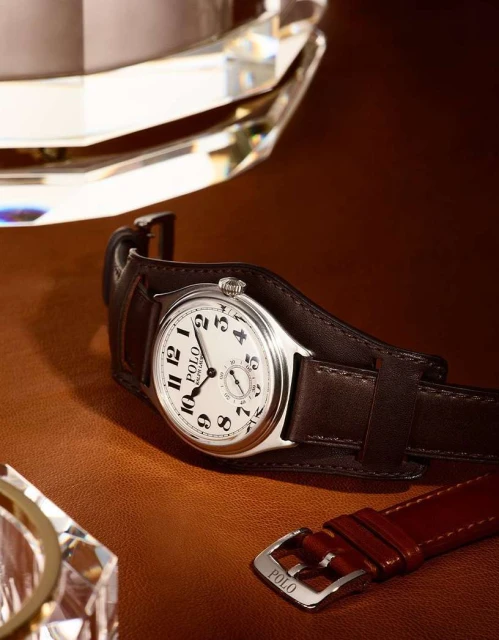 Silver and brown leather Polo Ralph Lauren watch on a brown surface next to a clear faceted crystal and leather strap detail.