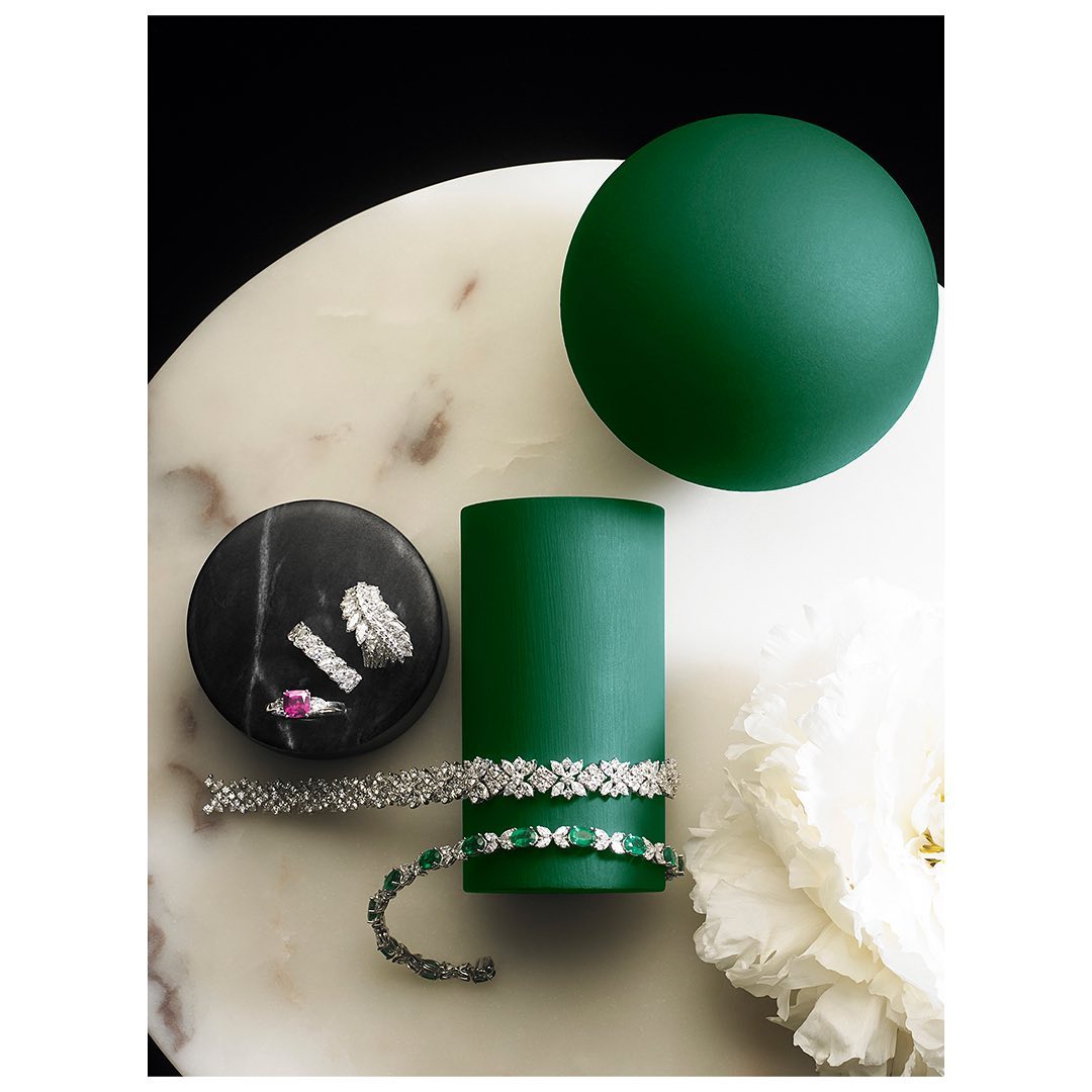Elevated still life of rings and bracelets displayed on a marble slab alongside a white peony and matte, jewel-toned geometric shapes.