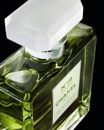 Close-up product shot of a rectangular glass perfume bottle with pale green liquid and a faceted white stopper against a black background
