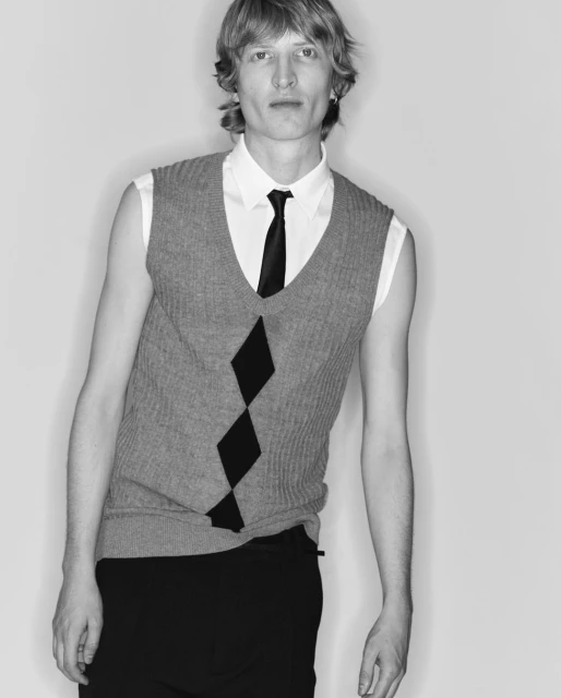 Monochrome portrait of a blonde male wearing a sleeveless sweater vest, white shirt, and black tie against a plain background.