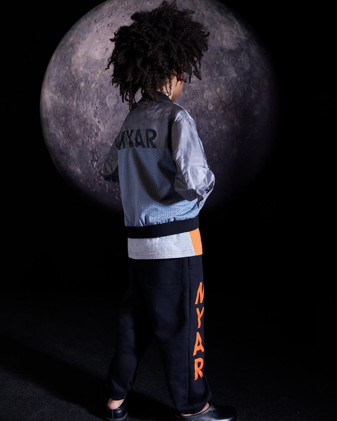 Young boy poses in a light jacket and dark pants with 'MYAR' text, set against a dark backdrop featuring a large moon.