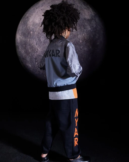 Young boy poses in a light jacket and dark pants with 'MYAR' text, set against a dark backdrop featuring a large moon.