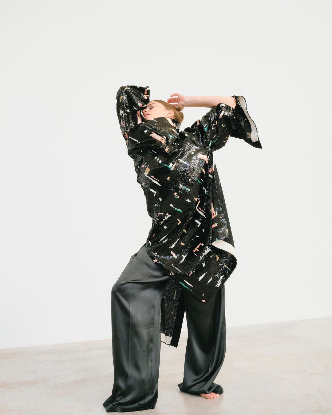 A person poses in a black patterned tunic and gray pants against a white backdrop in a fashion forward studio setting with dramatic lighting.