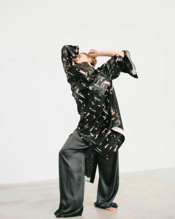 A person poses in a black patterned tunic and gray pants against a white backdrop in a fashion forward studio setting with dramatic lighting.