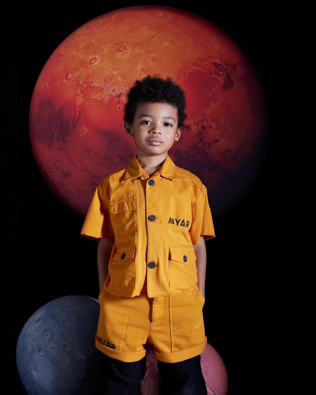 Young child poses in an orange MYAR matching short-sleeve button-down shirt and shorts against a dark space background with planets.