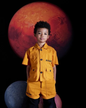Young child poses in an orange MYAR matching short-sleeve button-down shirt and shorts against a dark space background with planets.