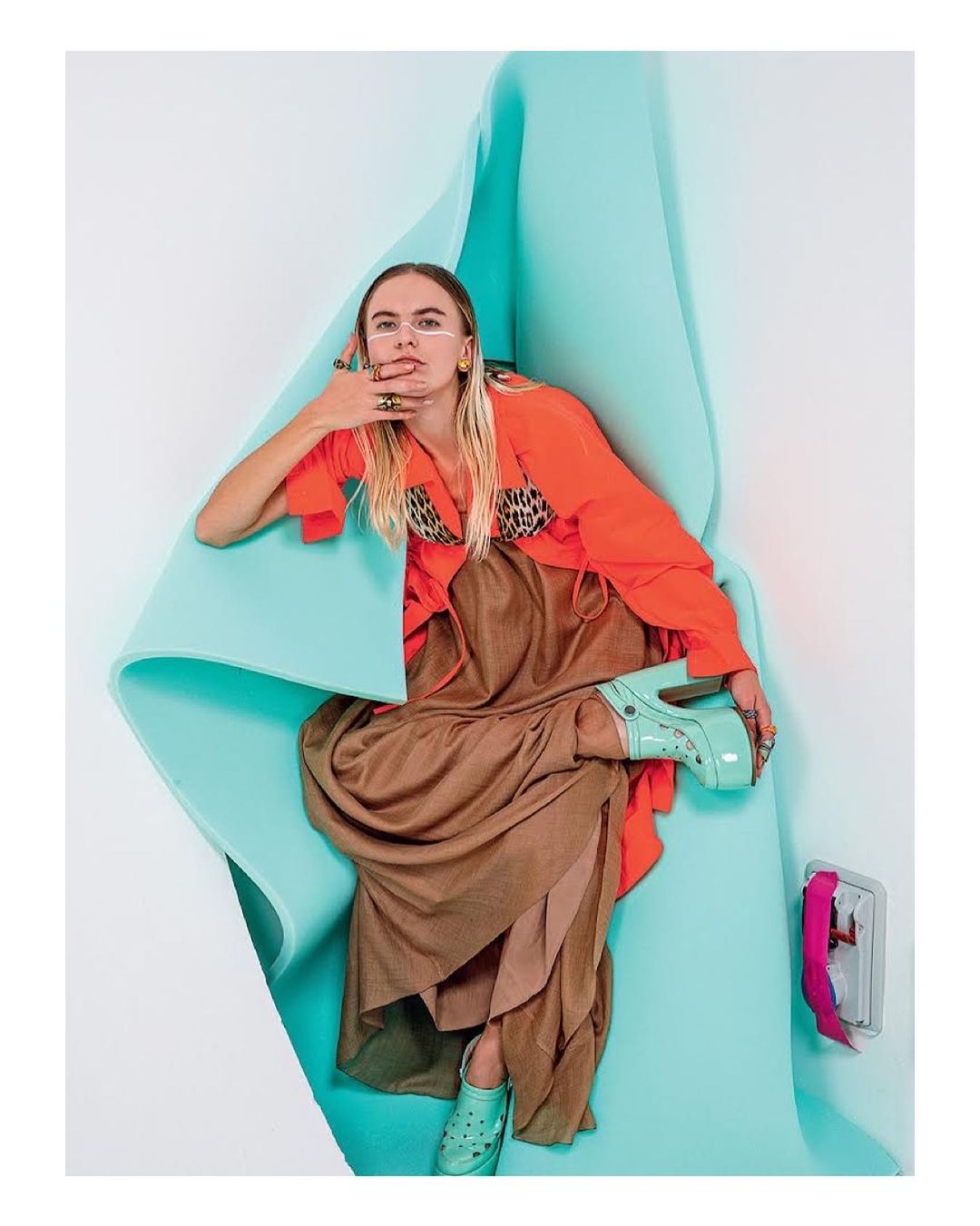 A woman in an orange top and long brown dress sits on a turquoise paper chair, with turquoise platform clogs, in a studio setting.