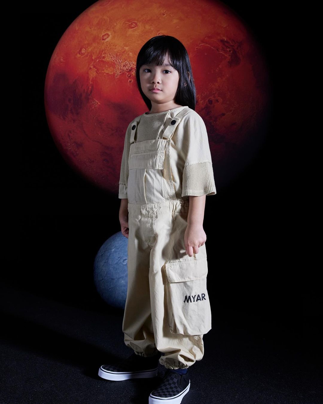 Child poses in beige overalls against a backdrop of Mars and a blue planet, accented by black checkered shoes in a studio setting.