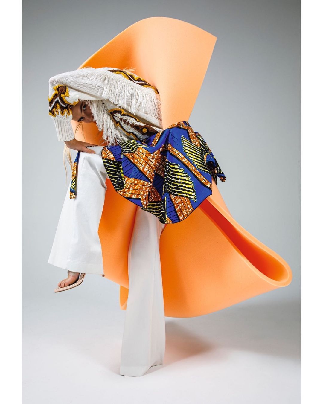 Avant-garde fashion shot features a model wearing a patterned blue and yellow garment, an orange sculptural piece, and white fringe.