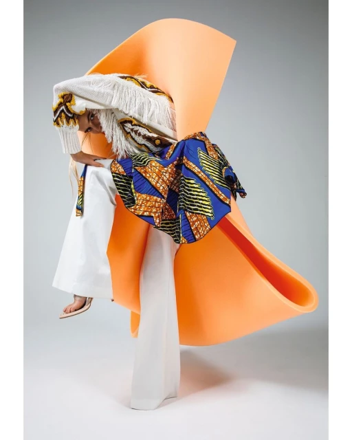 Avant-garde fashion shot features a model wearing a patterned blue and yellow garment, an orange sculptural piece, and white fringe.
