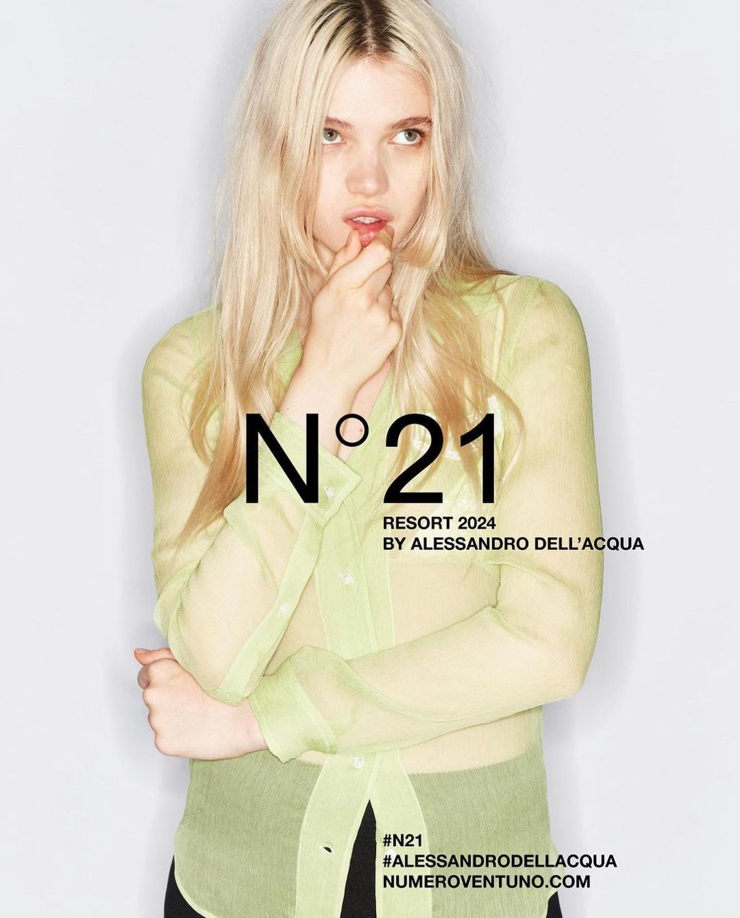Fashion shot of blonde adult female in a transparent button-down shirt in front of a white background, featuring N°21 brand text overlay.