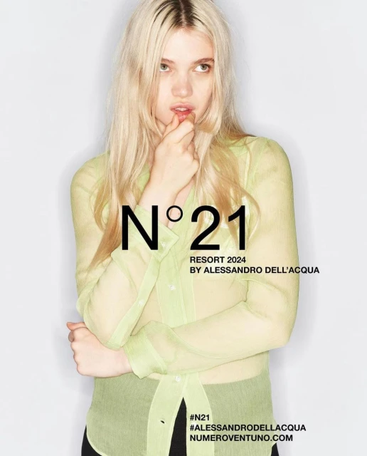 Fashion shot of blonde adult female in a transparent button-down shirt in front of a white background, featuring N°21 brand text overlay.