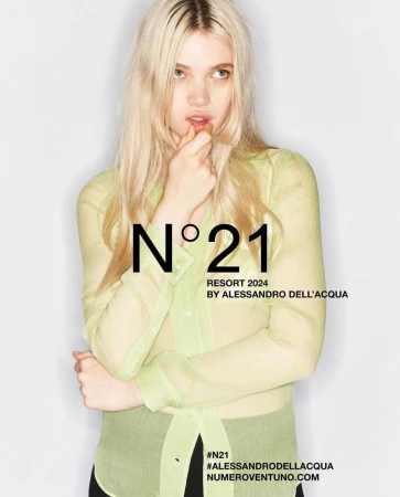 Fashion shot of blonde adult female in a transparent button-down shirt in front of a white background, featuring N°21 brand text overlay.