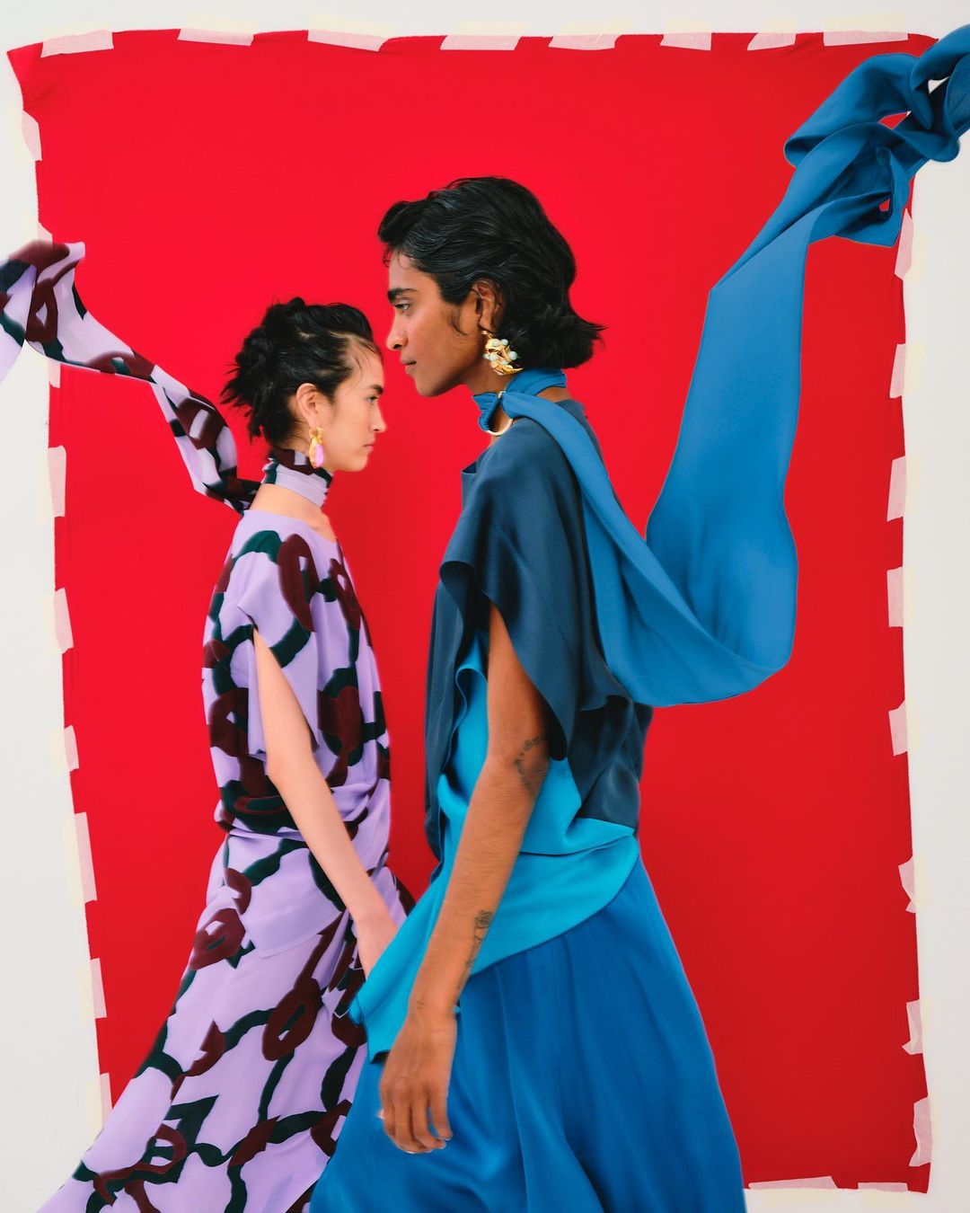 Two women face each other in a studio shot. One woman wears a lilac dress with dark brown pattern and the other wears a teal dress.