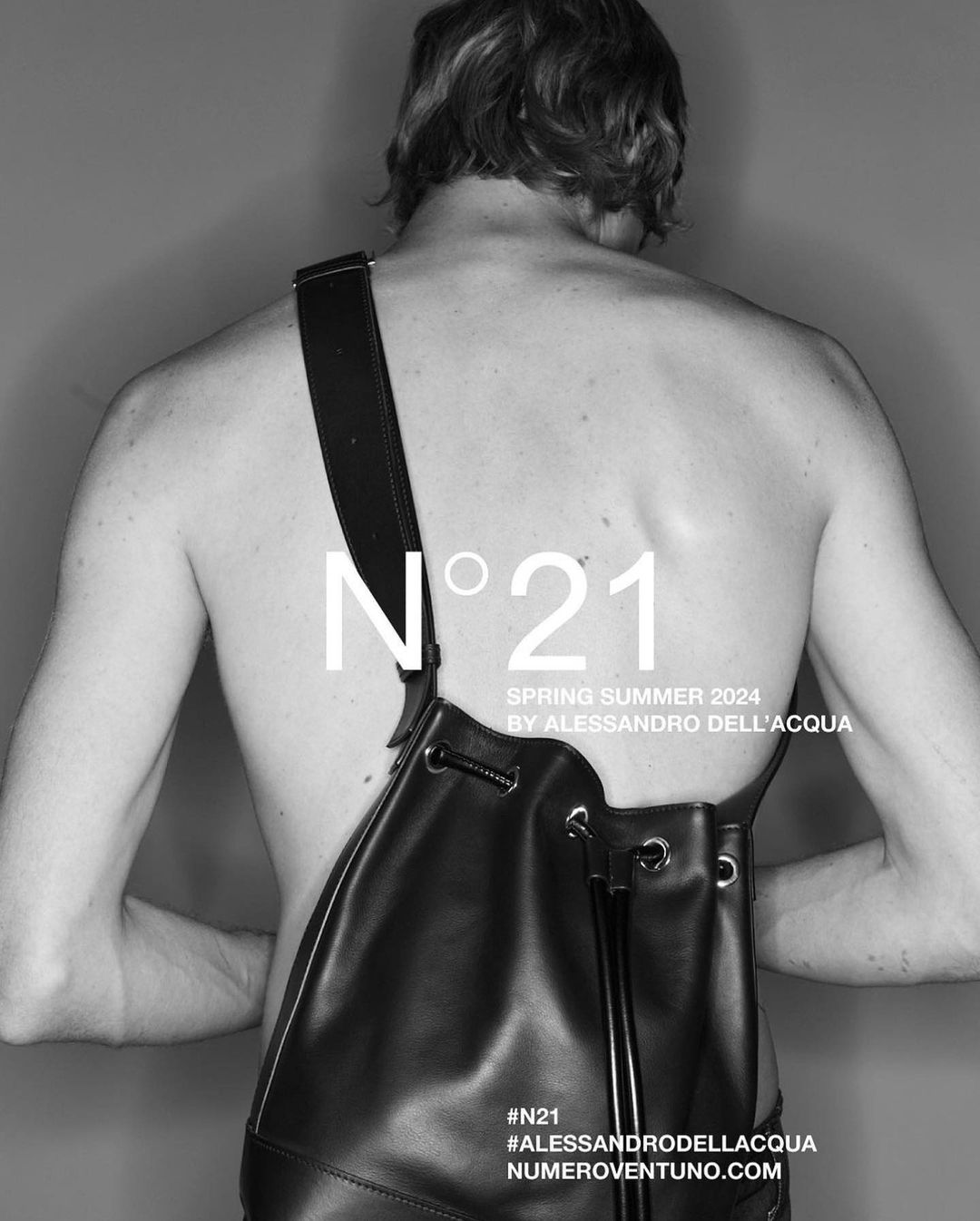 Black and white fashion ad featuring a shirtless adult male wearing a leather bag with N°21 brand logo and Spring Summer 2024 text.
