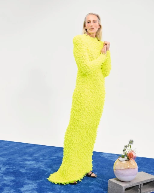 A woman poses in a long-sleeved, floor-length, bright yellow dress with a textured surface; a vase with flowers sits on a concrete block.