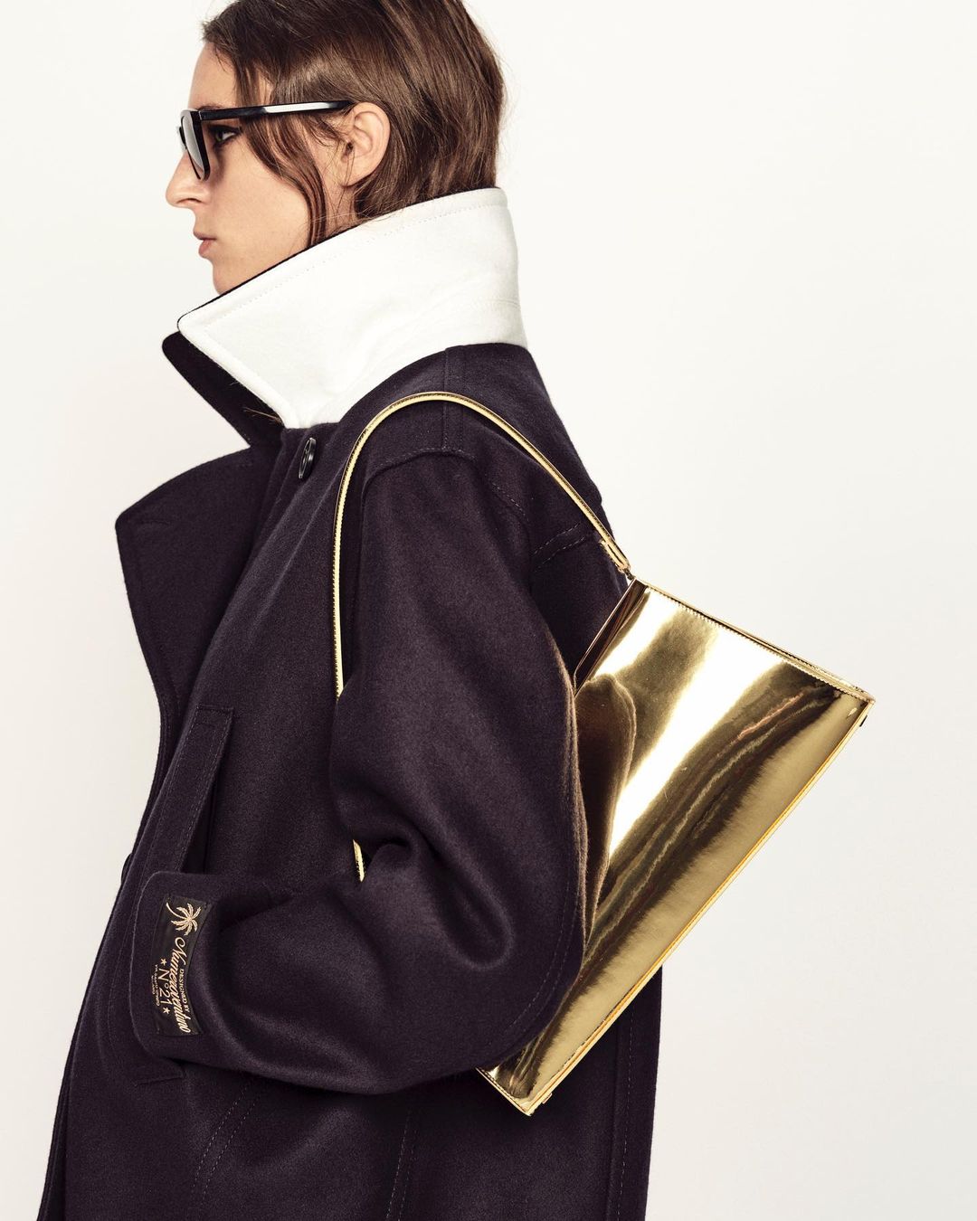 Fashion shot featuring a woman in a dark coat with a large white collar carrying a gold handbag, accessorized with sunglasses.