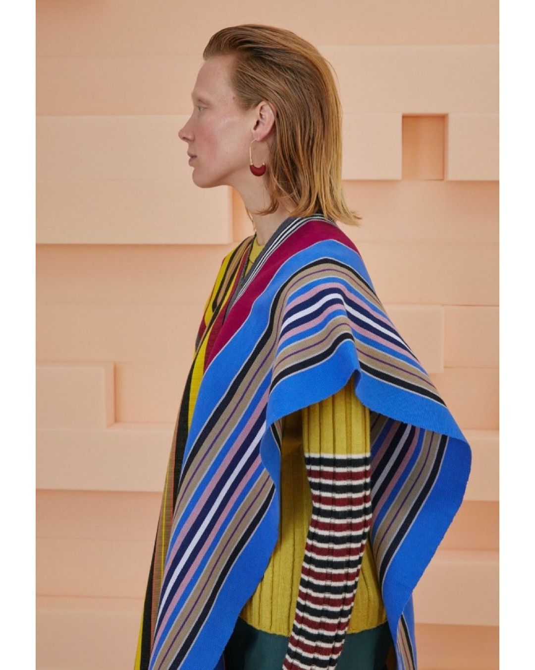 Side view of a stylish woman with short red hair wearing colorful striped poncho with earrings against a peach studio background.