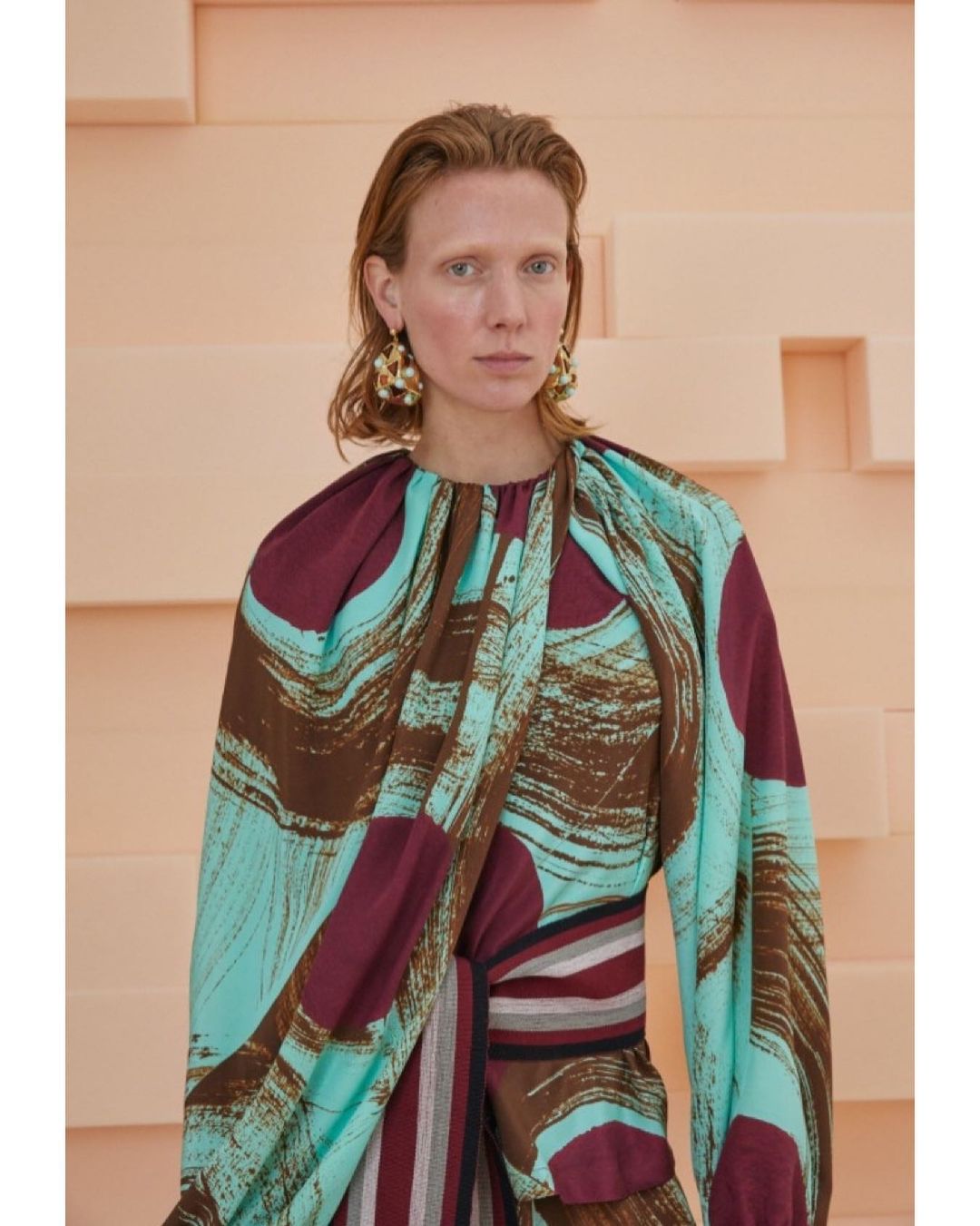 Adult female fashion model in a studio environment wears a burgundy, brown, and turquoise patterned dress with gold earrings.
