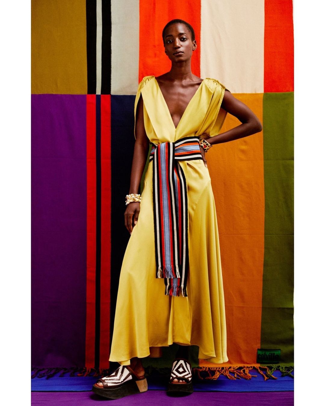 A dark-skinned model poses in a draped yellow dress and colorful striped sash against a backdrop of vertically striped textiles.