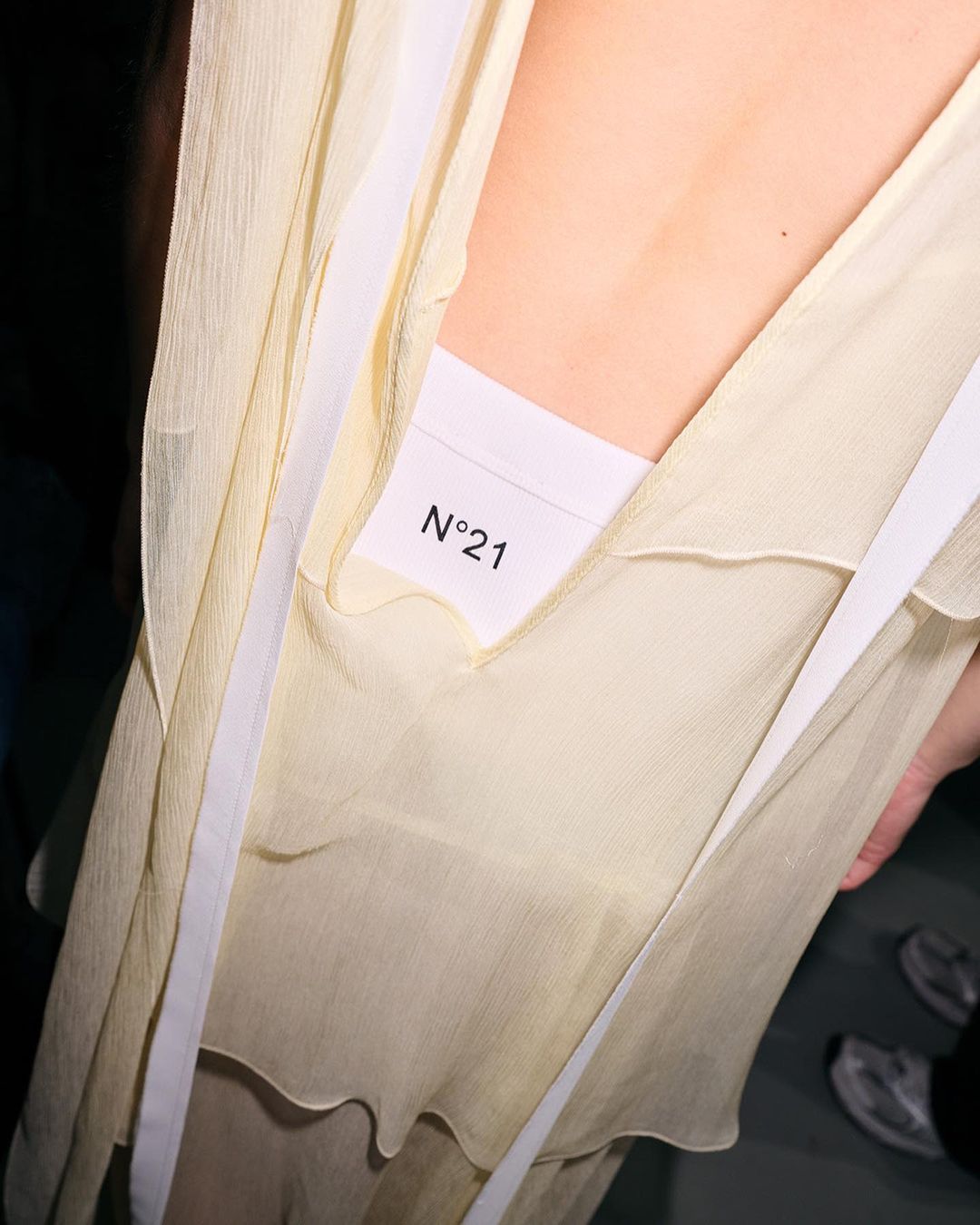 Close-up of a woman's back with light yellow, semi-sheer fabric and white ribbons, revealing a name tag with 'N°21' printed on it.
