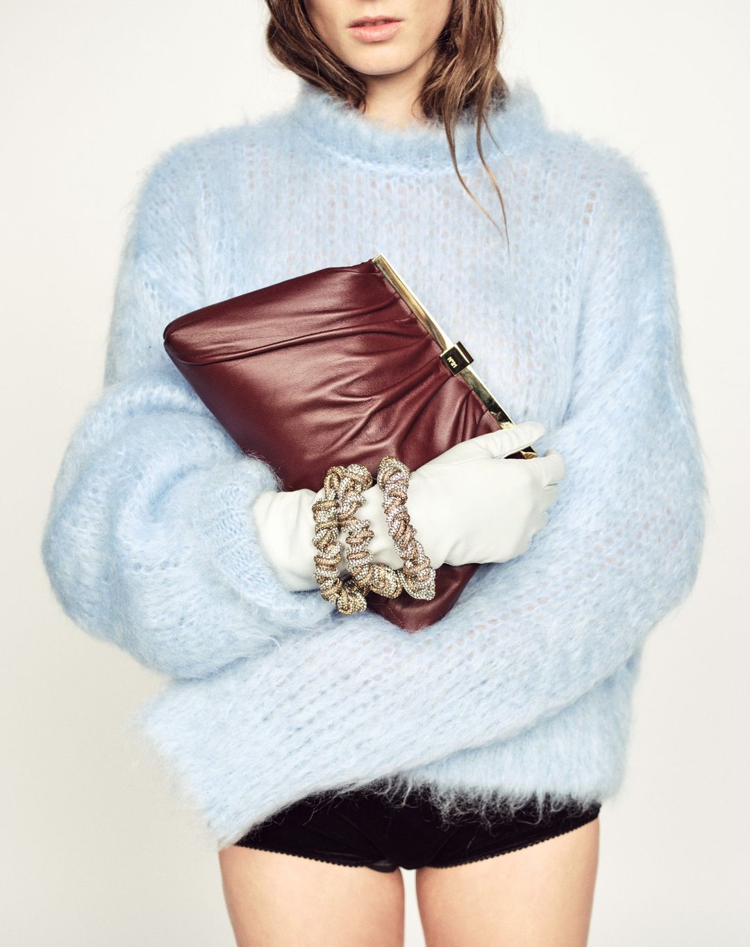 A woman in a fuzzy blue sweater and white gloves holds a burgundy clutch with gold hardware in a studio setting on a white background.
