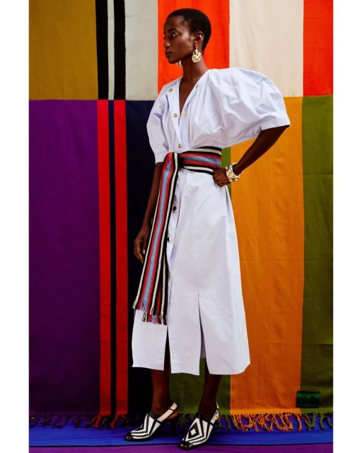 A Black woman in an off-white dress with a colorful belt poses against a multicolored patchwork backdrop, showcasing fashion and style.