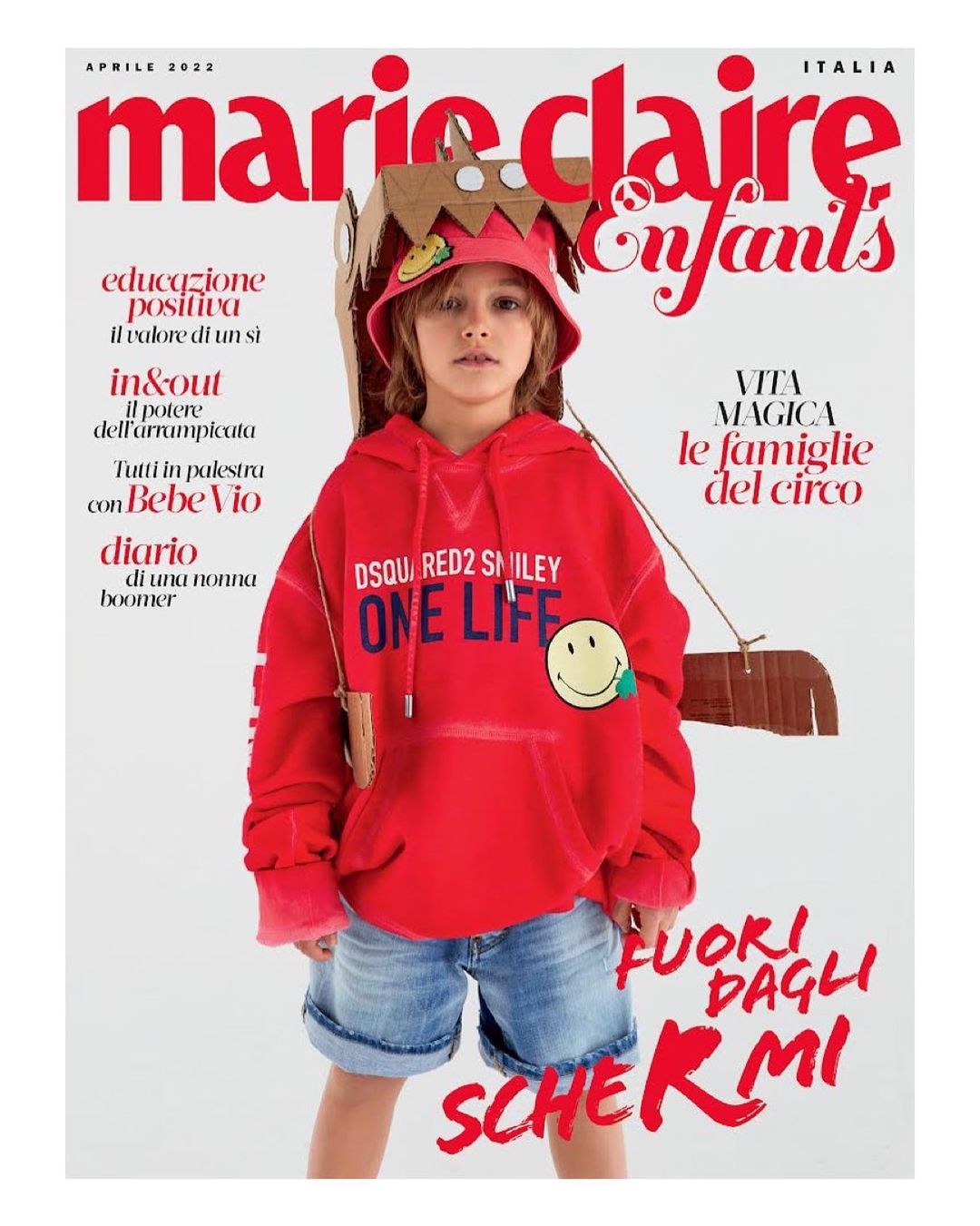 Magazine cover featuring a young child model wearing a red hoodie and denim shorts, styled with a cardboard hat and backpack against a white background.