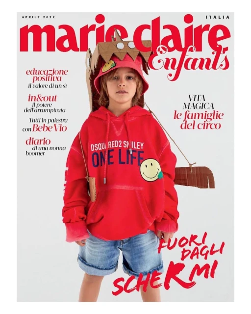 Magazine cover featuring a young child model wearing a red hoodie and denim shorts, styled with a cardboard hat and backpack against a white background.