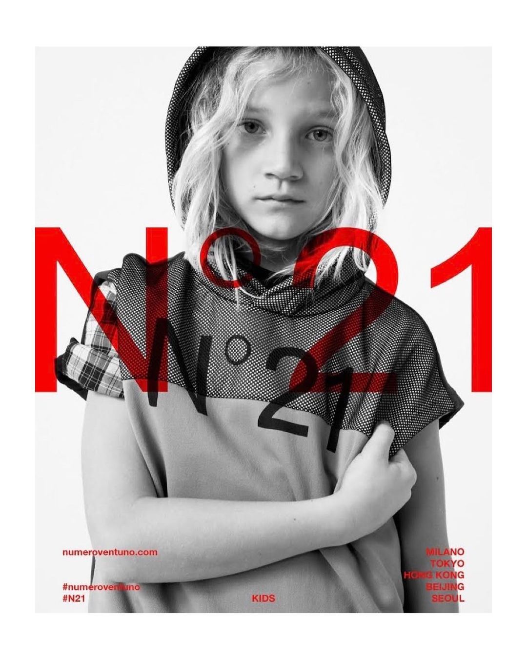 Fashion portrait of a child wearing a hooded top. Red 'N21' graphic overlay. White background, shot indoors. Fashion editorial, kids fashion.
