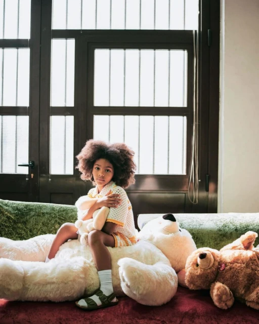 Young girl with afro hair sits on large teddy bear indoors by window wearing casual outfit with sandals and socks in a calm pose.
