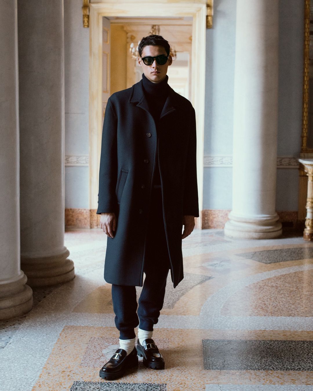 Man in black coat stands in a hallway with columns, wearing sunglasses, black turtleneck sweater, pants and shoes with white socks.
