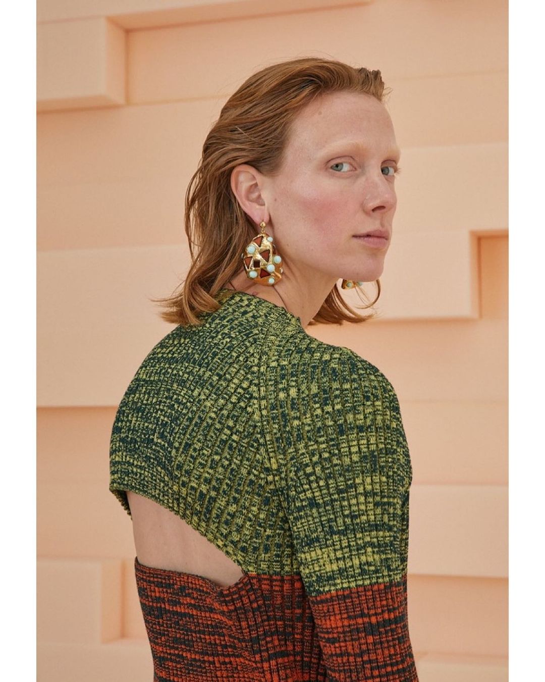 A fashionable adult female poses indoors with a textured peach wall background, wearing a stylish color-block knit dress and statement earrings.