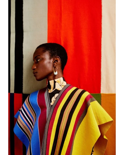 Elegant Black woman in a colorful striped poncho against a backdrop of red, black, cream, and green color blocks. Fashion editorial.