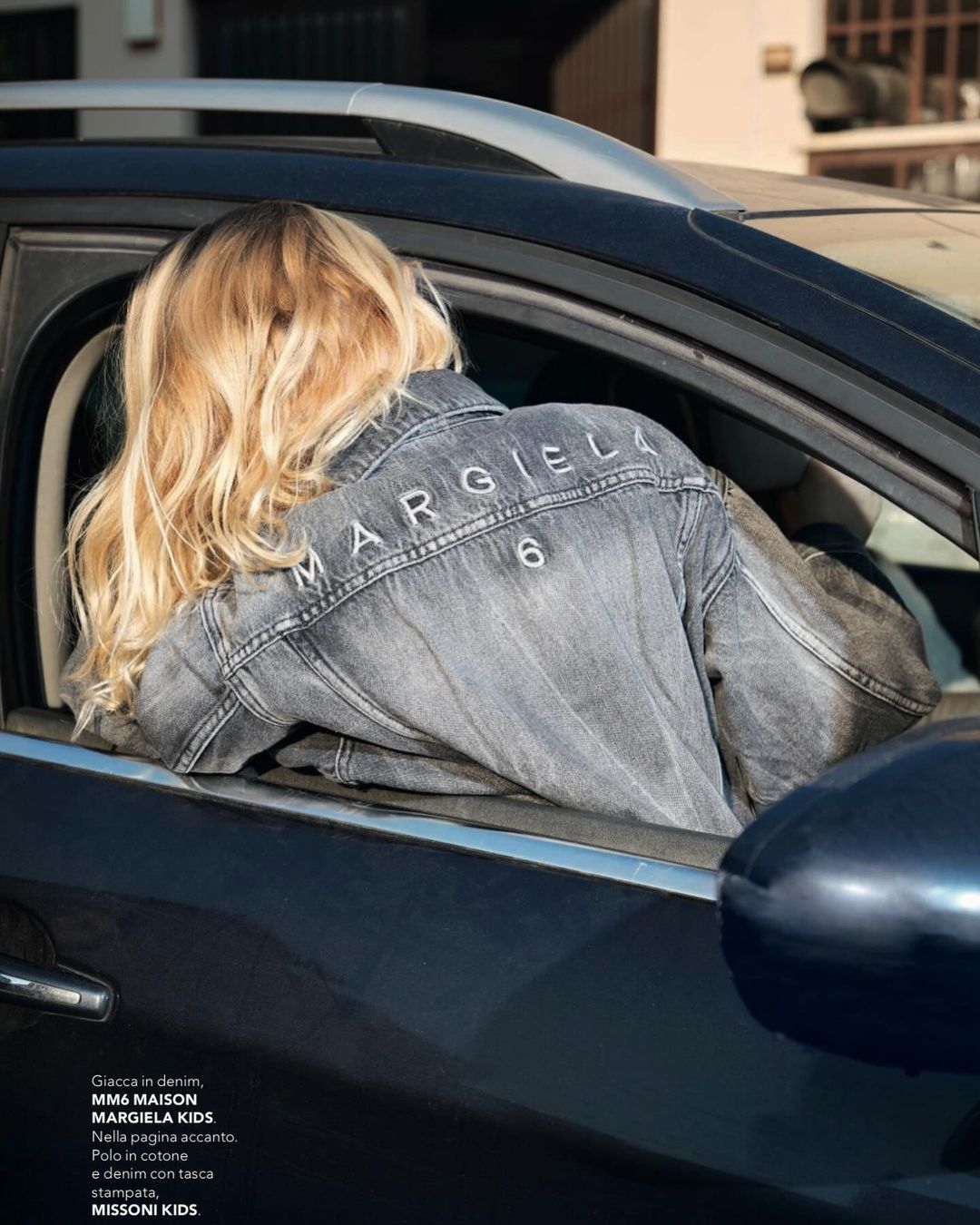 Blonde woman in a car wearing a denim jacket with Margiela logo. The focus is on the jacket and the car's interior.