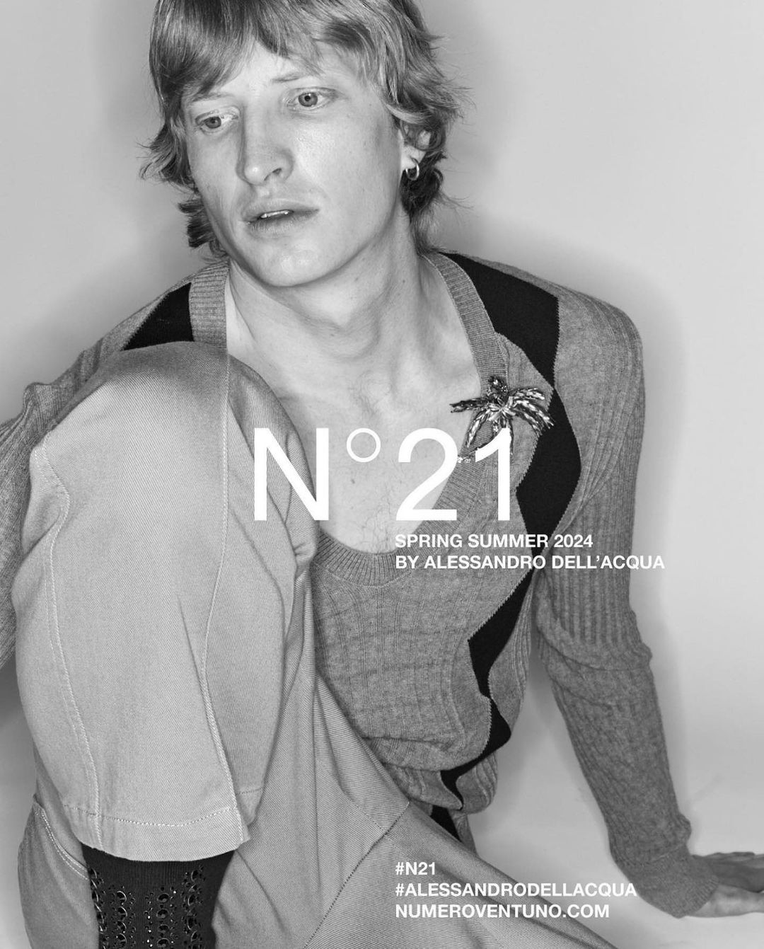 Black and white studio shot of a male model wearing a cardigan, grey trousers and an embellishment, posing in a studio.