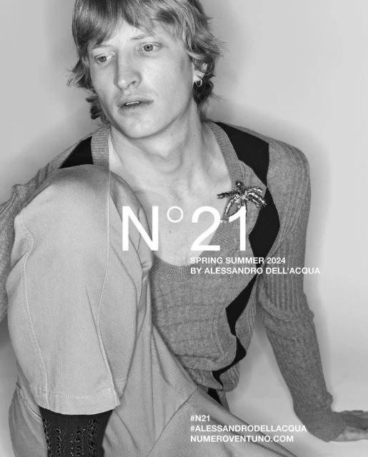 Black and white studio shot of a male model wearing a cardigan, grey trousers and an embellishment, posing in a studio.